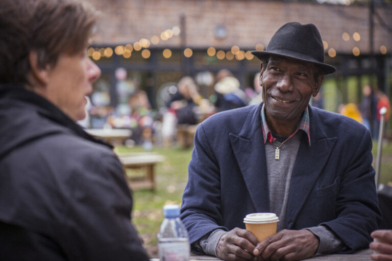 Older man with hat, smiling drinking a coffee outdoors at an event. Photo: Peter Kindersley. Source: Centre for Ageing Better