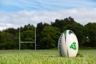 Ground level view of a rugby ball on a pitch, with the goal in the distance, in front of a row of dark green trees.
