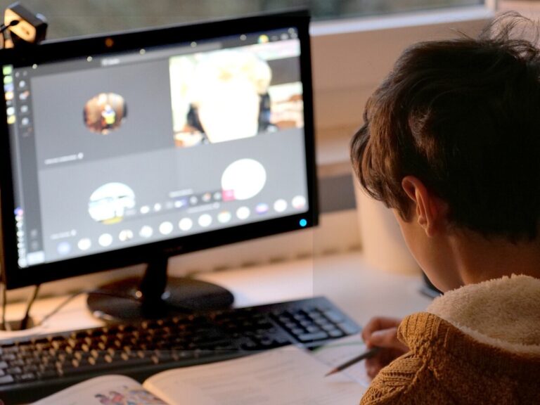 A young boy studies with exercise book and pen in front of his desktop computer