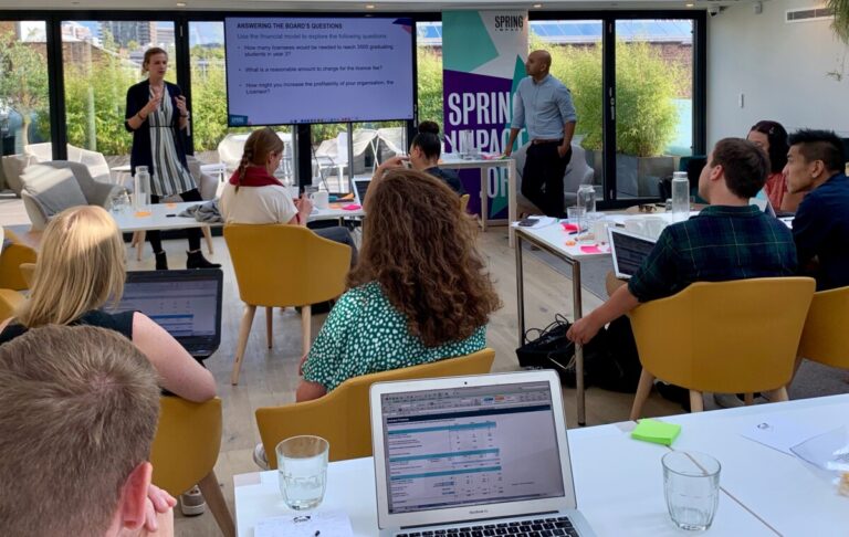 An audience of men and women sit in a modern office on mustard yellow chairs listening to a woman giving a speech by a powerpoint presentation on a board. She stands in front of a wall of windows, behind which are office blocks and trees