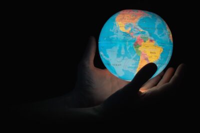 Against a dark background, a hand holds a small illuminated globe, showing north and south america