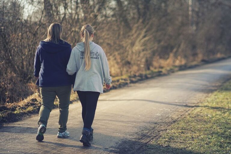 A woman and her daughter walk away from the camera down a path with trees on one side and grass on the other, bathed in sunlight