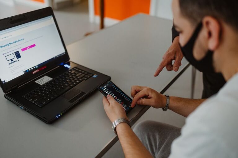 A man in a black mask looking at his phone, in front of a laptop