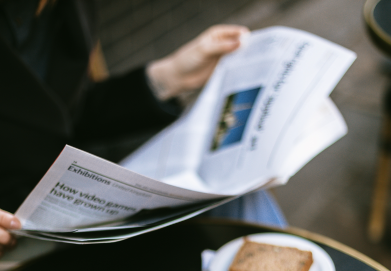 someone reading a newspaper outside a cafe with a slice of cake on a white plate