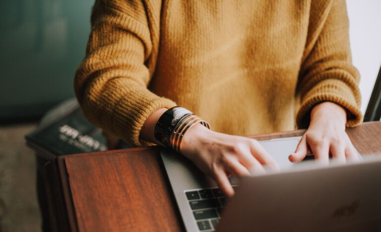 A woman in a mustard coloured sweater types on a laptop, on a wooden desk