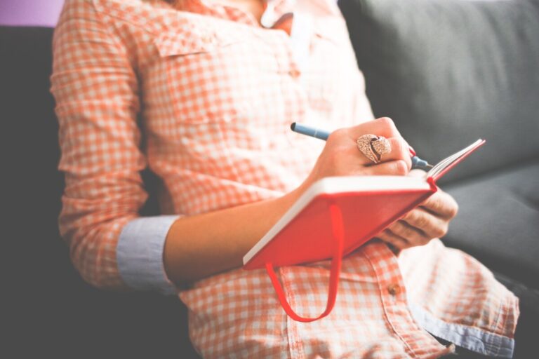 a woman in a red and white checked shirt writes notes in a red notebook
