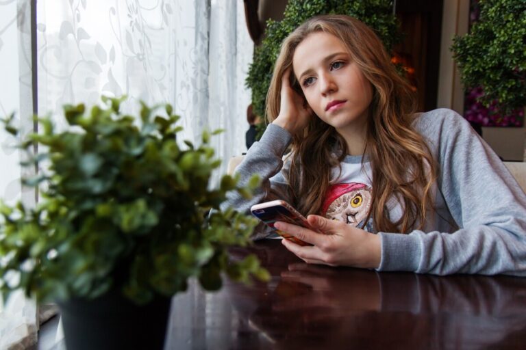 a girl gazes out of a window, holding her phone in one hand, and resting her head on the other