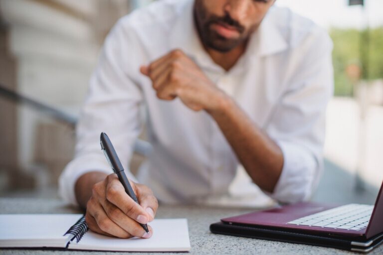 A man in a white shirt takes notes by his laptop