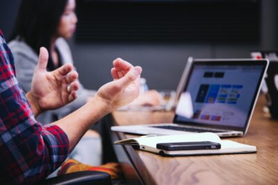 two people sit at a laptop. you can see the arms of a man in a plaid shirt who looks like he is explaining something to the woman