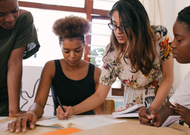 Women gather around a desk. One of them stands as she writes notes as the others pay close attention. Photo by RF._.studio on Pexels