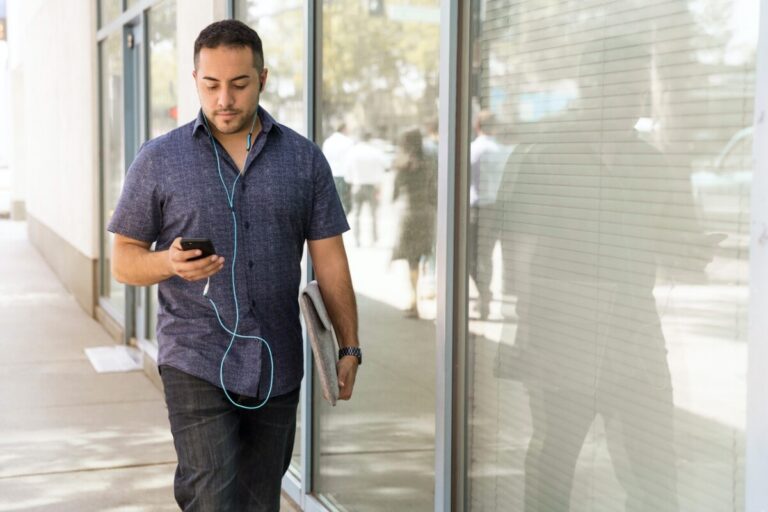 Man in tshirt walking down a sunny street, listening to a podcast on his phone. Image: Pexels.com