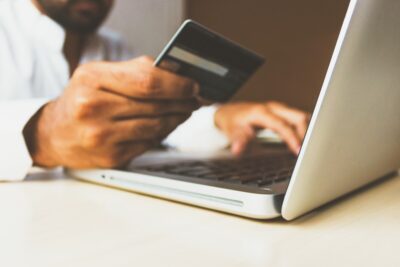 A man sits at his laptop, holding his debit card. By Rupixen on Unsplash