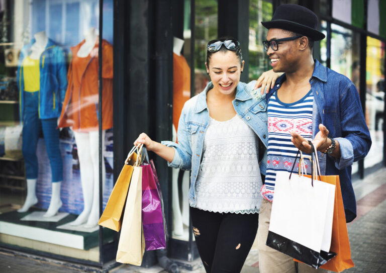 A smiling couple walk past a shop carrying bags of purchases