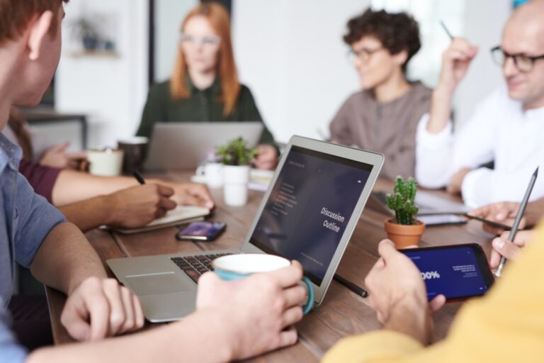 People sit around a table with an open laptop showing a document entitled discussion outline. By Fauxels on Pexels