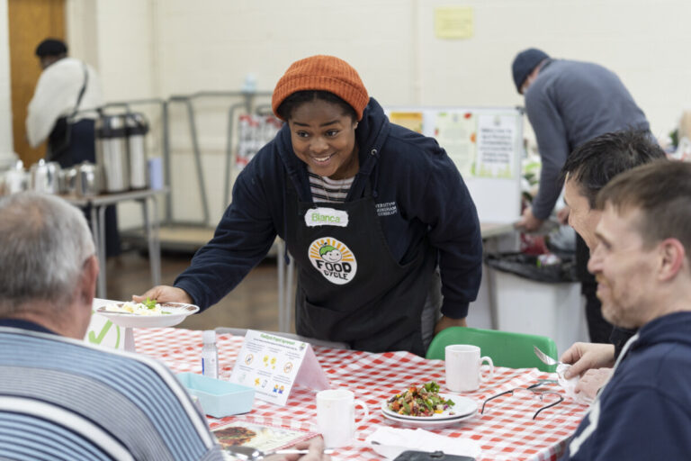 A woman in a Foodcycle t-shirt hands out a meal at a table of guests