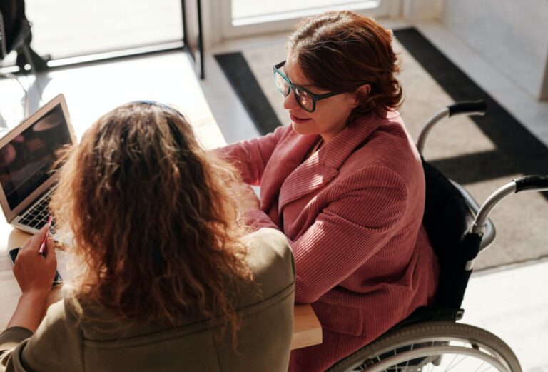 Two women (one in a wheelchair) sit at a table by a computer, talking. By Marcus Aurelius on Pexels