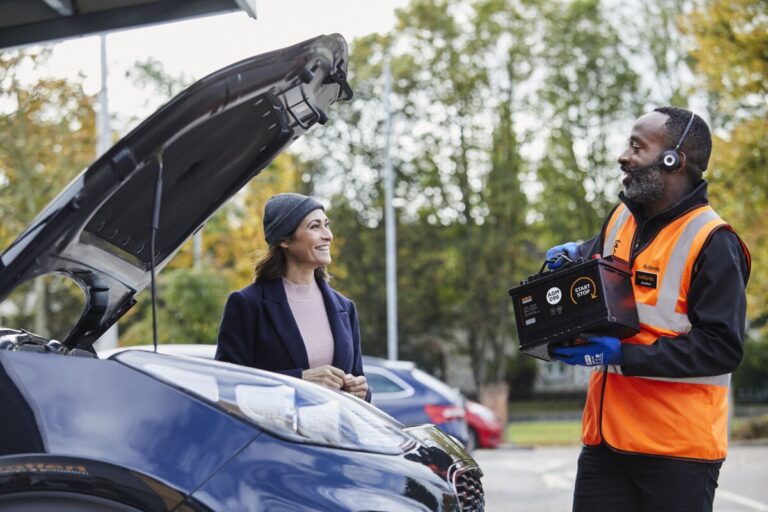 A mechanic brings a battery over to a woman standing by her car, which has the bonnet open