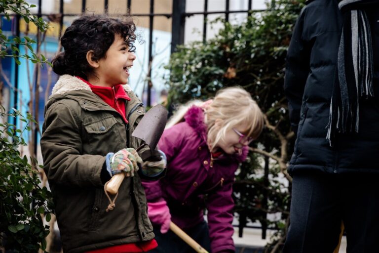 A child outside in a garden laughs as she holds a trowel alongside another child digging