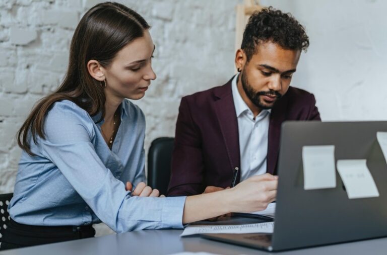 a man and a woman study some figures in front of a laptop in an office. The laptop has post-it notes on the back and the office walls are white painted brick. By olia danilevich on Pexels