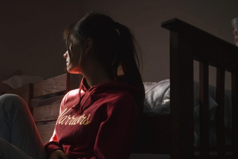 A woman leans against a bed and gazes into the distance. By Sofia Alejandra on Pexels