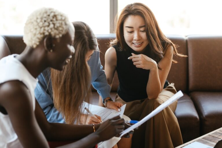 three women smile whilst looking at paperwork in a meeting. By Alexander Suhorucov on Pexels