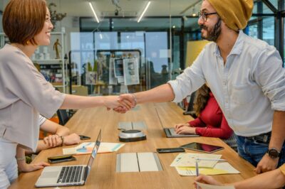 A man and a woman smile as they shake hands over a long wooden table. By Ketut Subiyanto on Pexels