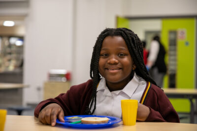 A school girl with a bagel and drink smiles at the camera