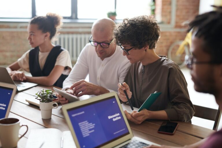 A bald-headed man in dark-rimmed glasses and a white shirt talks to a woman with short curly brown hair in a brown top in a meeting. By Fauxels on Pexels