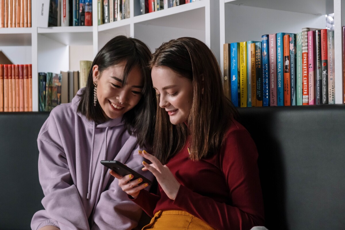 Two young women smile as they look down at a phone. By Cottonbro on Pexels