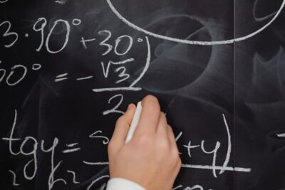 A hand draws maths equations on a blackboard in white chalk