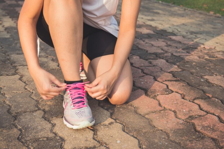 a woman in black shorts and white t shirt kneels and ties the pink lace on her trainer. By Tirachard Kumtanom on Pexels