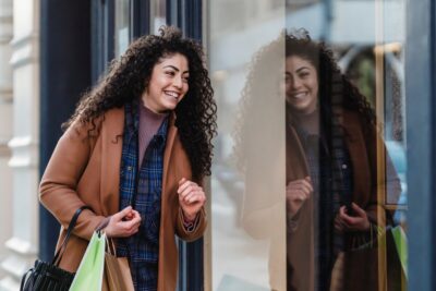 A woman with shopping bags smiles as she passes a shop window. By Tim Douglas on Pexels