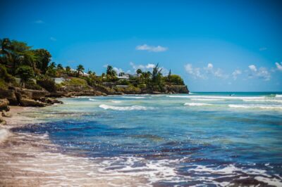 A picture of the blue ocean with the beach and rocky landscape behind it in Barbados. By Tom Jur on Unsplash.