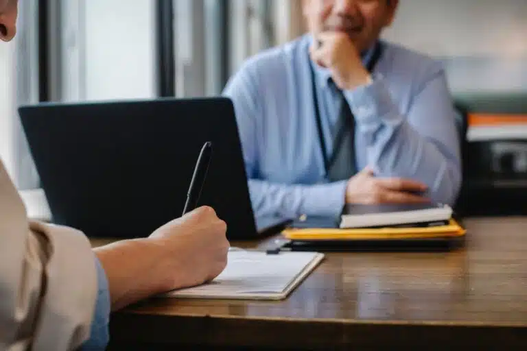 Two people sit across each other at a desk. A man in a blue shirt and tie listens and waits as a woman's hand writes with a pen on paper. By Sora Shimazaki on Pexels