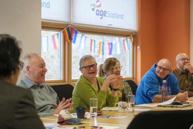 A group of older people sit talking and laughing around a long table in front of a window with bunting across it.