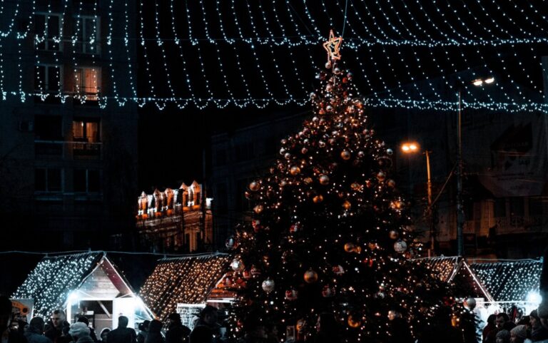 People gather around a Christmas tree in a town centre in the dark, illuminated by christmas and building lights. By Sebi Pintilie on Pexels