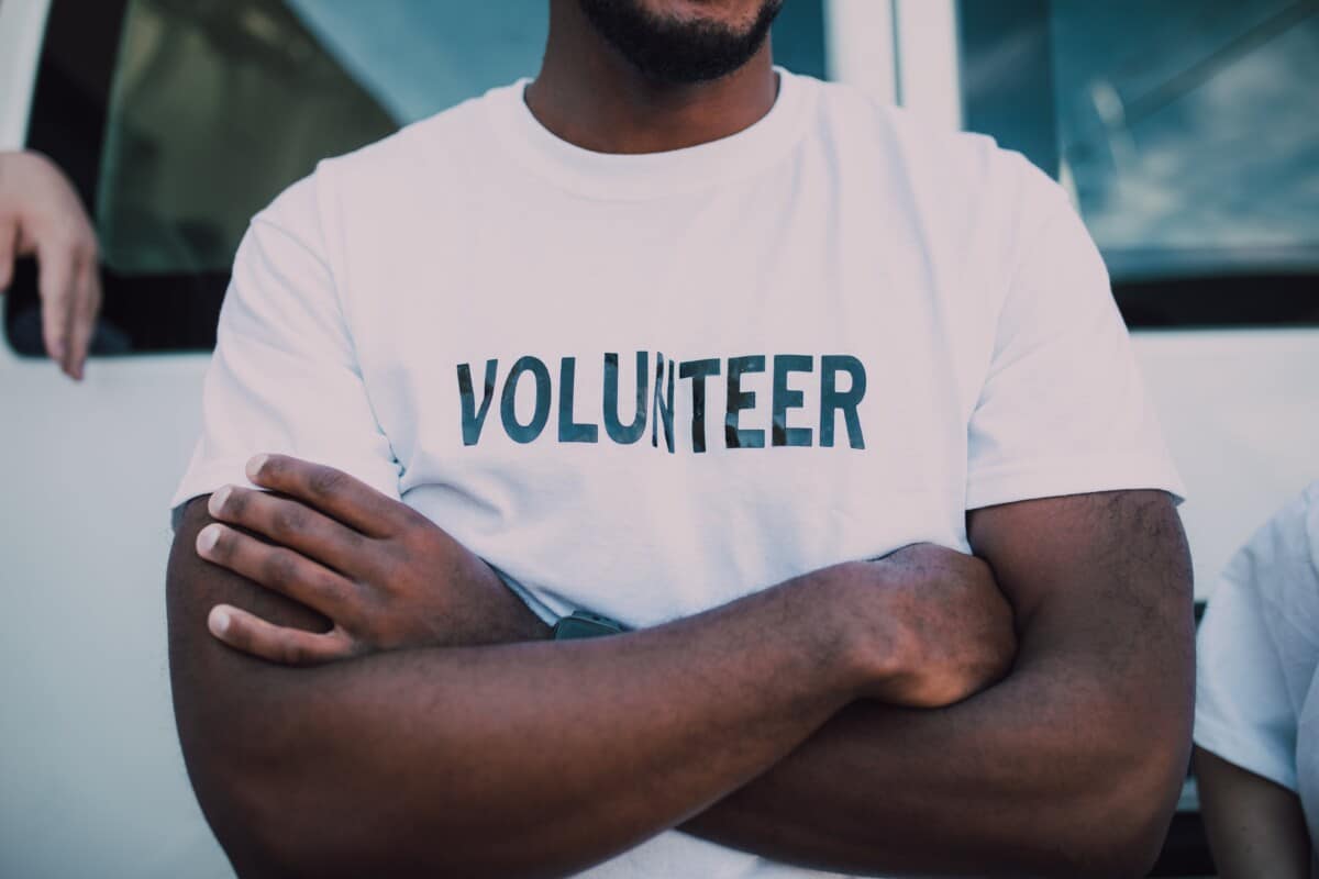 A man with his arms folded, wearing a white t-shirt with the word volunteer on it. By RDNE Stock Project on Pexels
