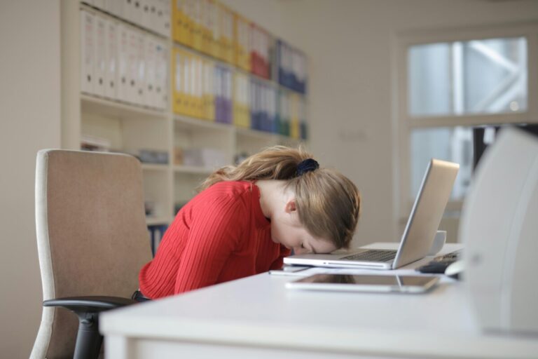 A woman sits with her head on her keyboard in an office room. By Andrea Piacquadio on Pexels