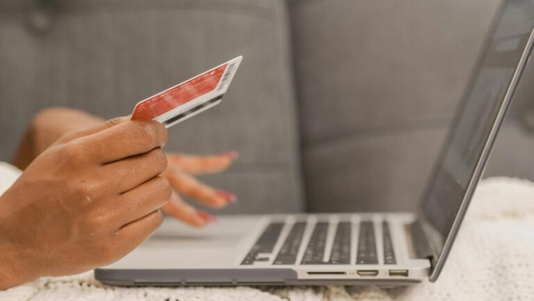 A hand holds a card while the other types on a laptop keyboard, making a payment. By Kindel Media on pexels