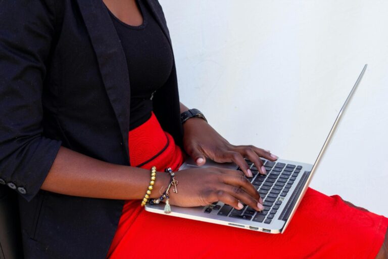 A woman in red and black types on a laptop that sits on her lap. By Oudney Patsika on pexels