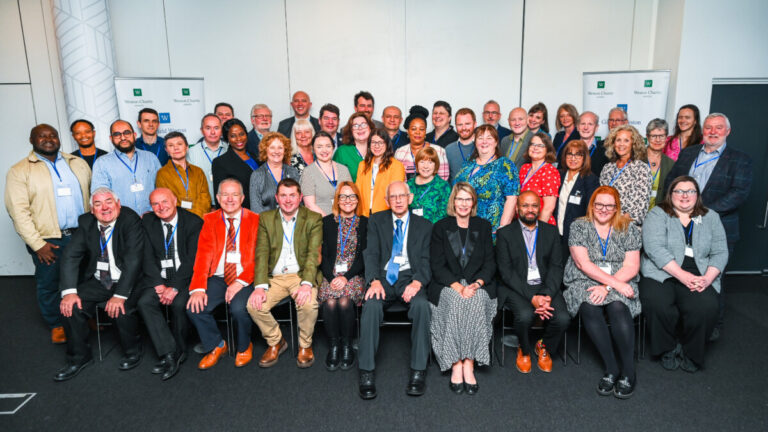 A large group of people, some seated and some standing, pose together, smiling and facing the camera. They are in front of signs for the Weston Charity Awards, and are all from this year's winning charities.