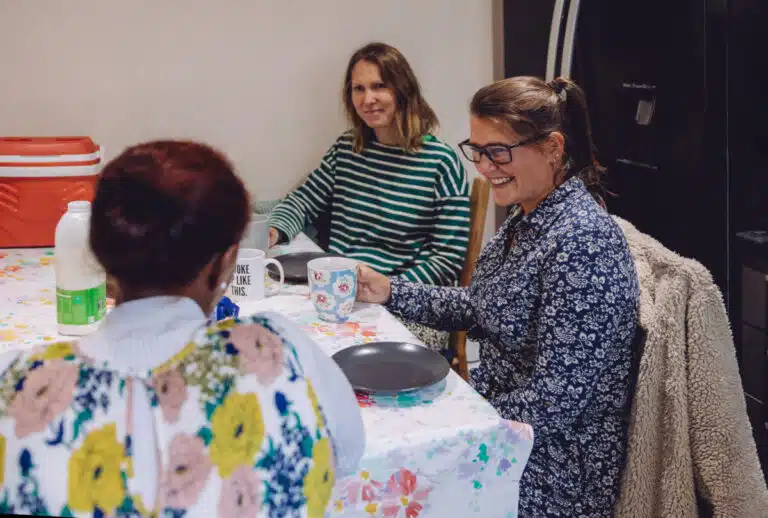 Three women sit around table chatting with plates and mugs. They are from the charity Ella's house