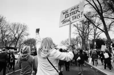 A black and white photo of people protesting with a woman holding a placard reading 'we are better than this!' By Stocksnap on Pixabay