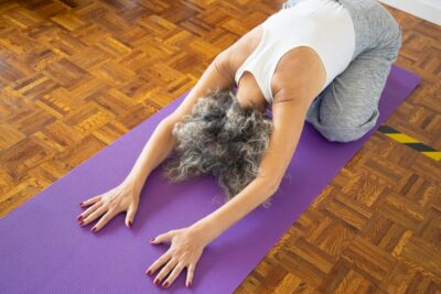 A woman with grey hair stretches over a purple exercise mat. By Kampus Productions on Pexels/