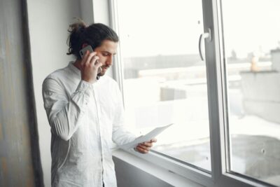 A man stands in front of a window, on the phone and lookin down at a sheet of paper