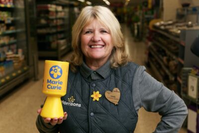 Morrisons staff member Linda smiles as she holds a Marie Curie collection box, and wears a daffodil pin