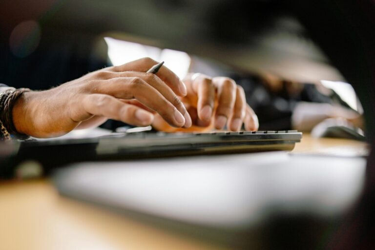 Hands typing on a keyboard, with a pen between two fingers. By Yan Krukov on pexels