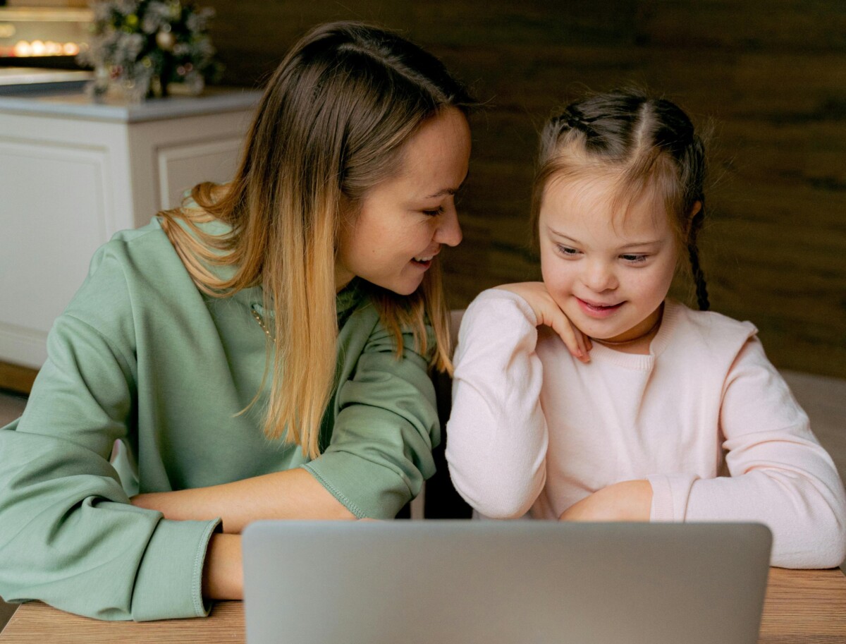 A mum and her daughter at a table with a laptop in front of them. The daughter is a young girl with down Syndrome, wearing a light pink top and with her long brown hair plaited. Her mum is wearing a light green top and looking at her daughter, smiling. By Antoni Shkraba