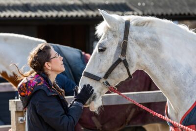 A woman in a wheelchair, with her hair in a plait, and wearing glasses, looks up at a whote horse, as she strokes its nose. From Riding for the Disabled.