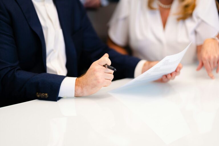 A man and a woman in an office sit at a table. The man is holding a sheet of paper and a pen. By Carrie Allen on Unsplash.